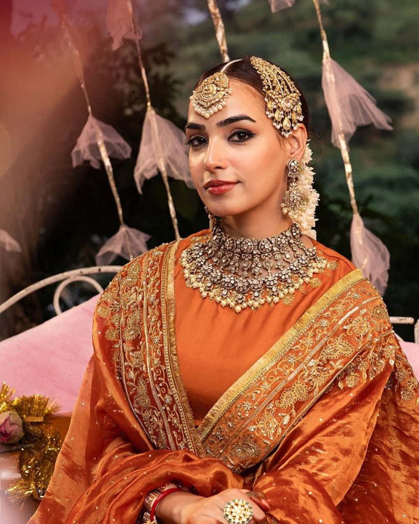 Bride in orange ensemble wearing heavy polki choker with layered pearl drops and matha patti. 