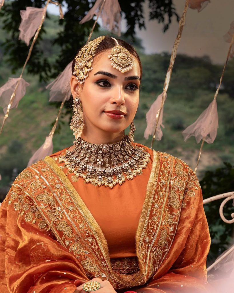 Bride in orange ensemble wearing heavy polki choker with layered pearl drops and matha patti. 