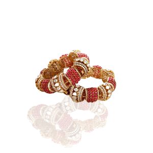 Ornate gold bangles with ruby beads and polki diamonds by MJR Jewels placed on a beige surface with soft pink flowers.