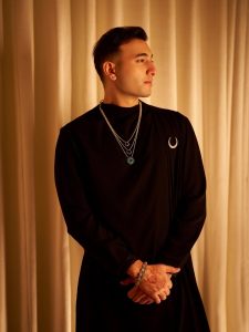 Male model in black kurta wearing layered delicate chain necklaces with gemstone pendants.