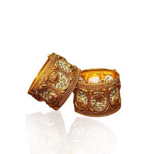 Intricately carved gold bangles with green accents by Rakesh Jewellers placed on a warm golden surface.