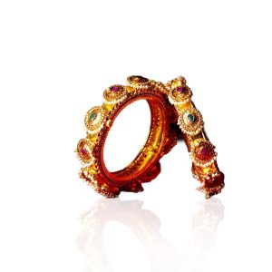 Temple style gold bangles with pearl and gemstone detailing by Rakesh Jewellers on a dramatic red background.