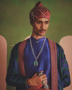 Male model in traditional attire wearing chunky silver chain necklace with emerald pendant and statement ring.
