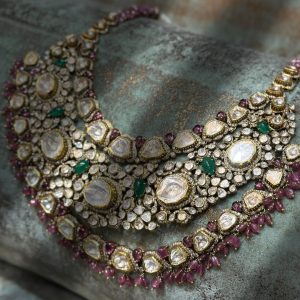 Three layered polki necklace with emerald and ruby accents by Shree Jee Jaipur placed on a textured stone surface.