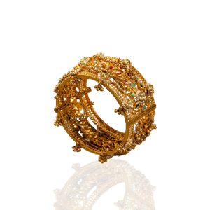 Detailed gold filigree kada with pearl accents by Suvarnam Jewels on a textured neutral background.