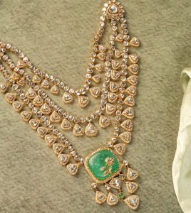 Polki necklace with triangular diamond motifs and a large emerald centre pendant by Suvarnam Jewels on a soft green background.