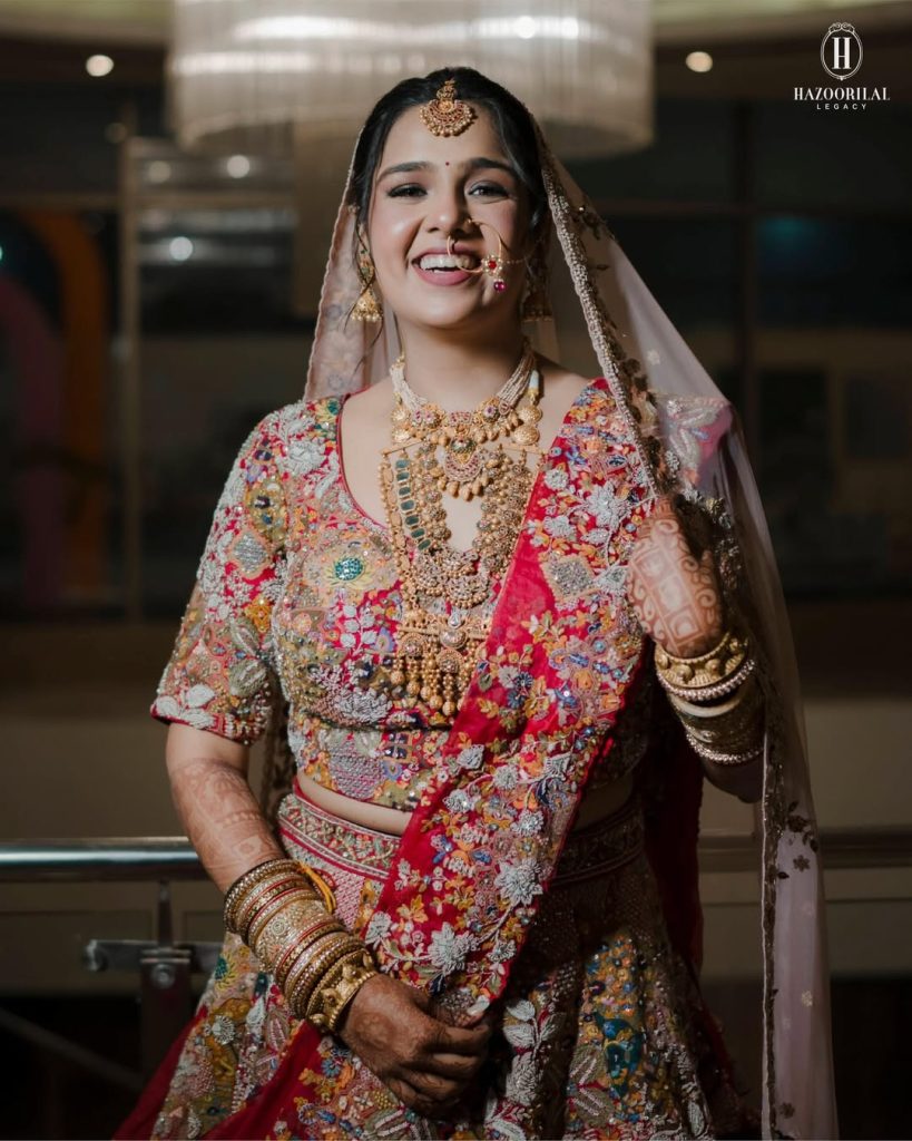 Bride in multicolour lehenga wearing layered gold necklaces with coloured gemstone detailing, traditional nath and stacked bangles