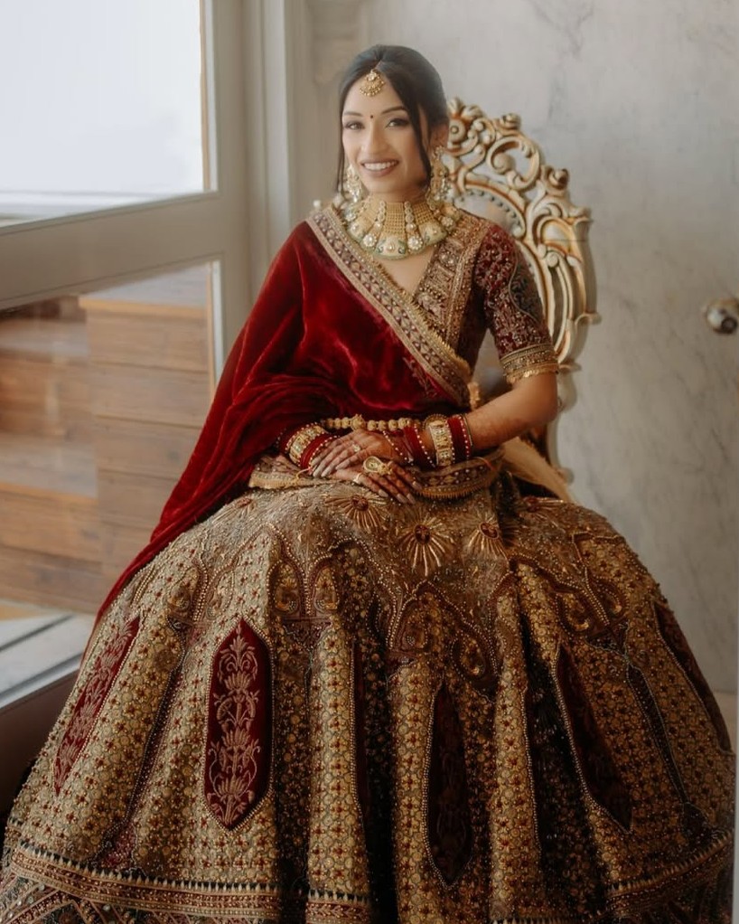 Bride in maroon velvet lehenga with gold choker, green and white stone detailing, kamarband and traditional bridal jewellery
