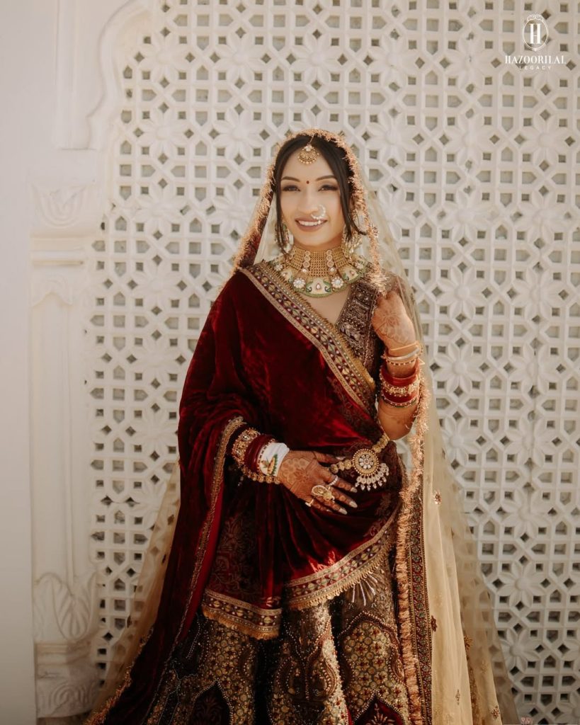 Bride in maroon velvet lehenga with gold choker, green and white stone detailing, kamarband and traditional bridal jewellery